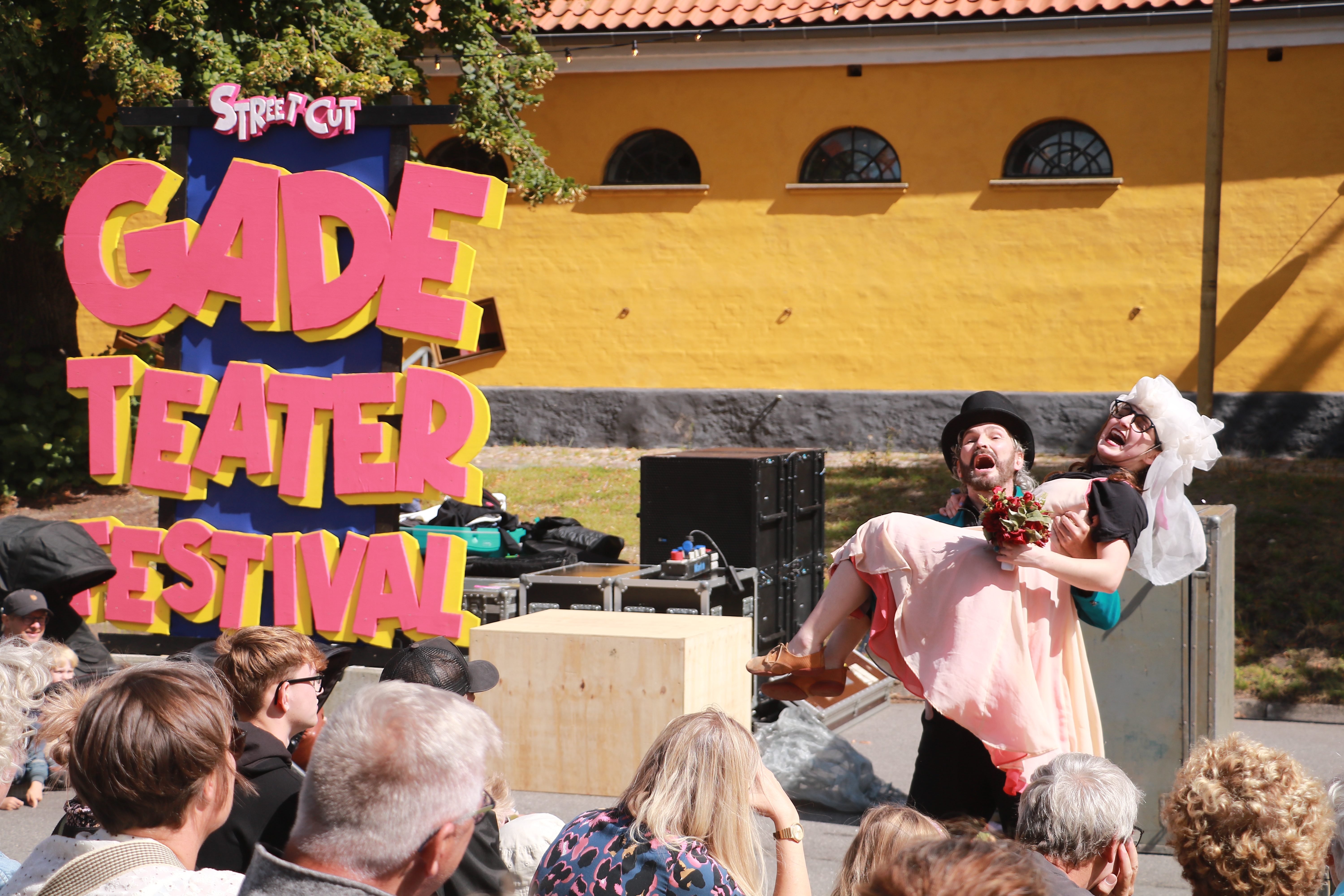 Stort skilt, hvor der står Steet Cut Festival i venstre side. To skuespillere klædt ud i slør og høj sort hat. Den ene bærer den anden liggende i armene. Publikumshoveder i forgrunden. 