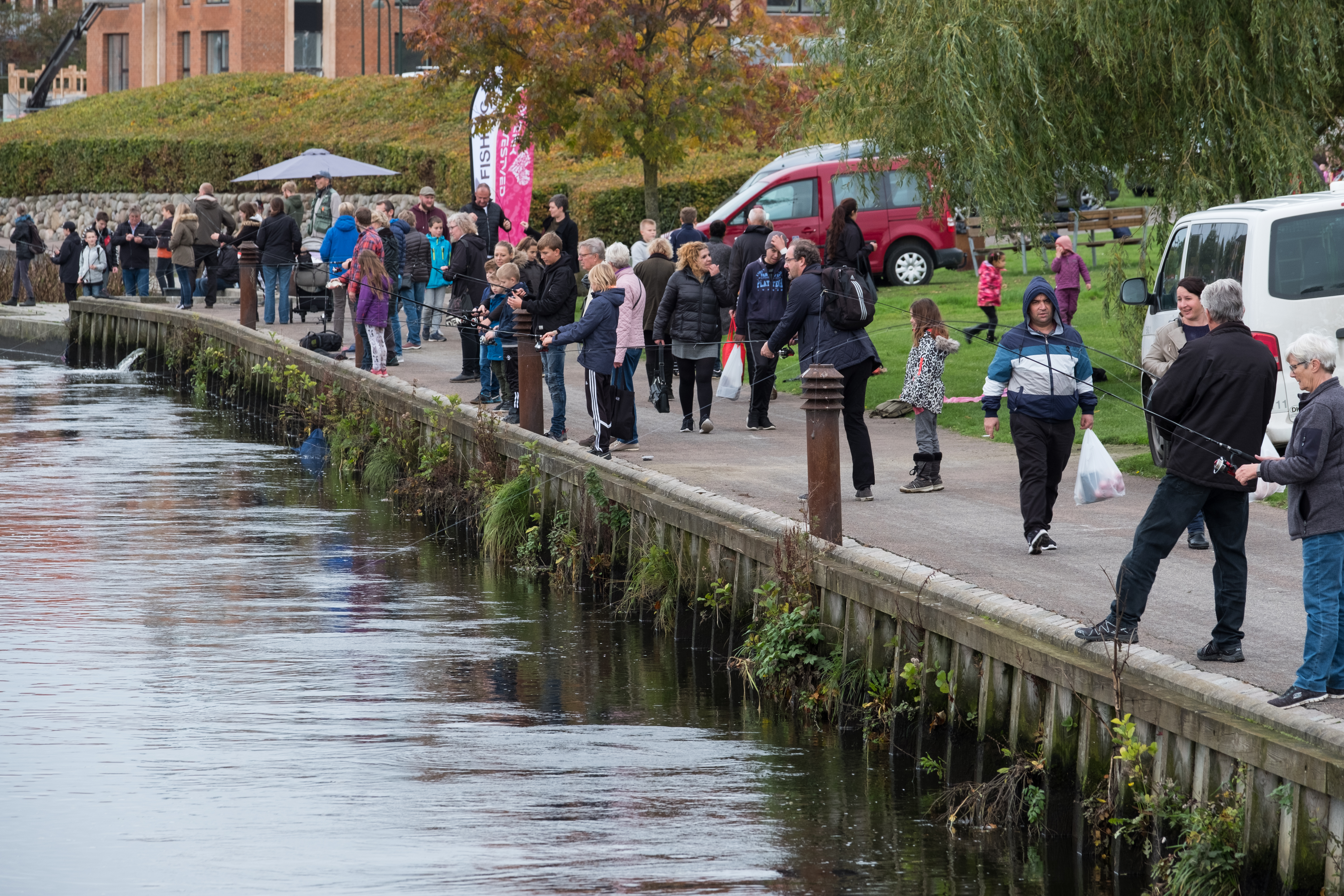 Børn, unge og voksne står og fisker langs en å. 