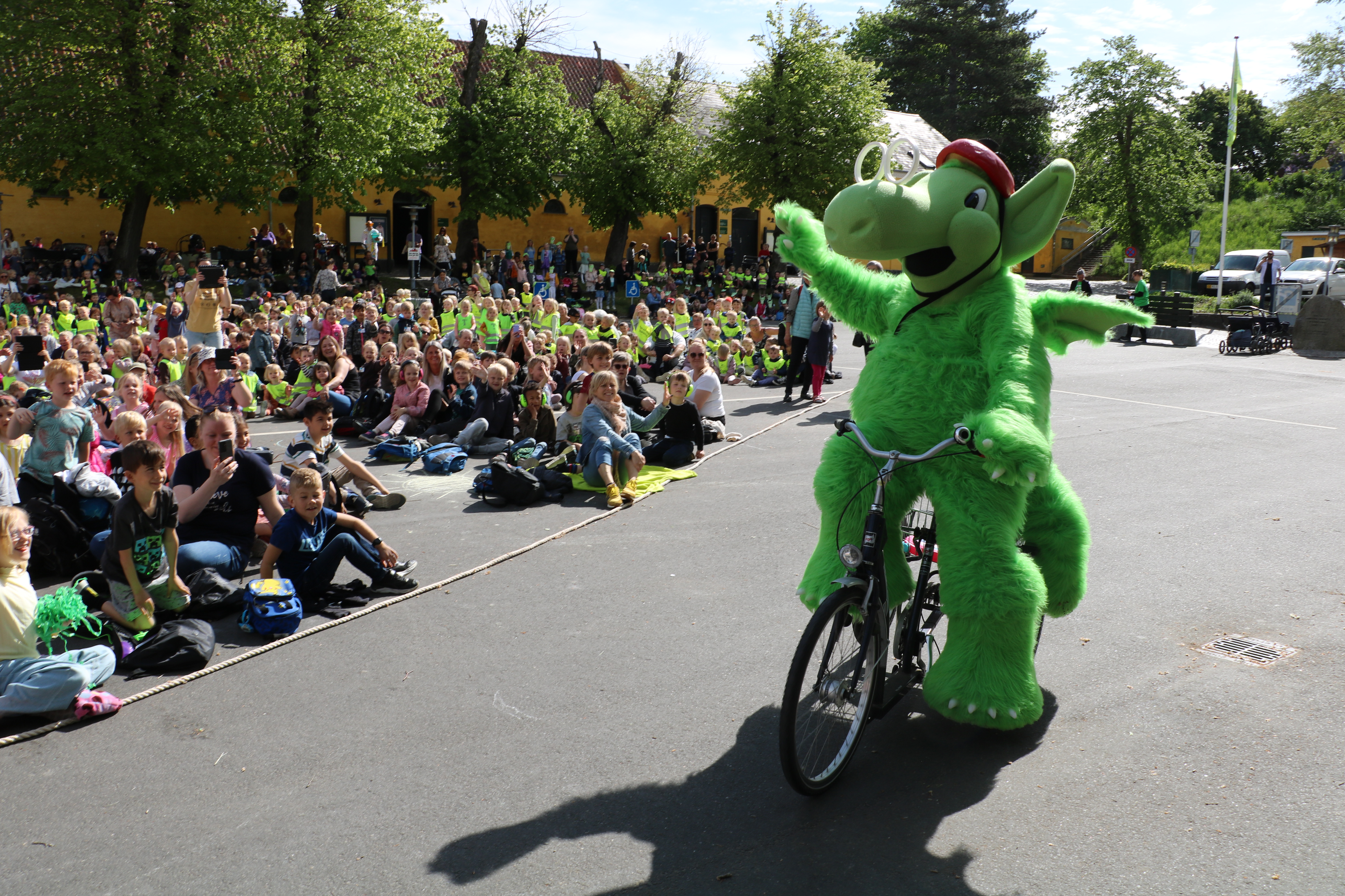 Stor, grøn plysdrage, Næssi, på cykel foran en masse børn til børnekulturfestival på Grønnegade Kasernes Kulturcenter. 