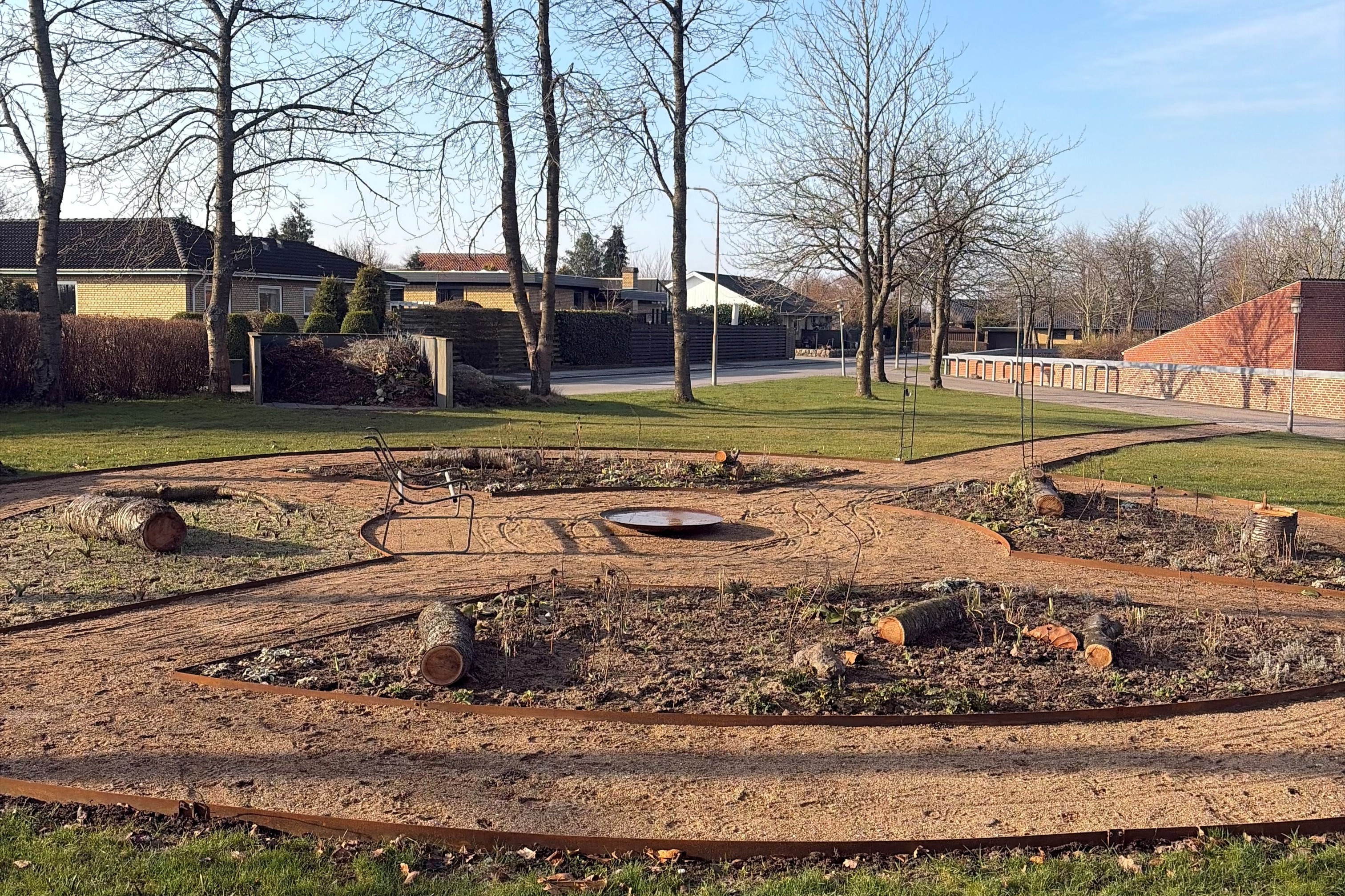 Foto af de nyanlagte havebede med planter og træstubbe, en bænk og et vandfad.