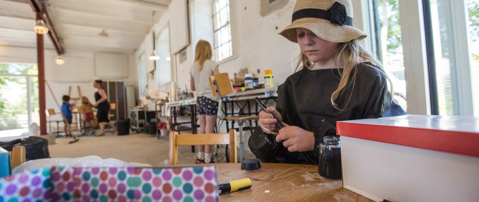 En lille pige med hat, er ved at tage hætten af en tusch imens pædagoger og børn er i baggrunden