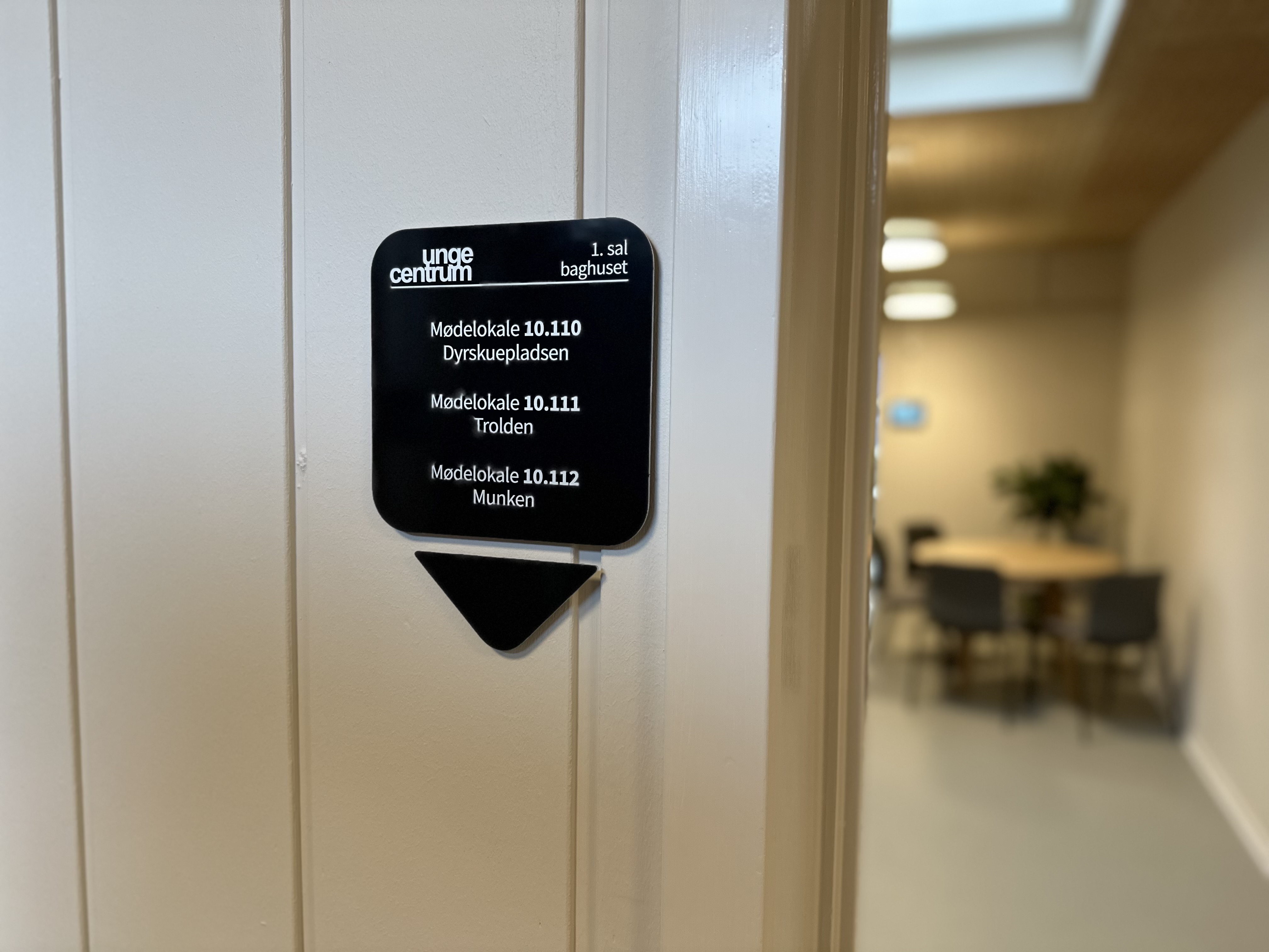 Skilt i forgrunden med navne på mødelokalerne: Dyreskuepladsen, Trolden og Munken. Bord med stole i baggrunden. 