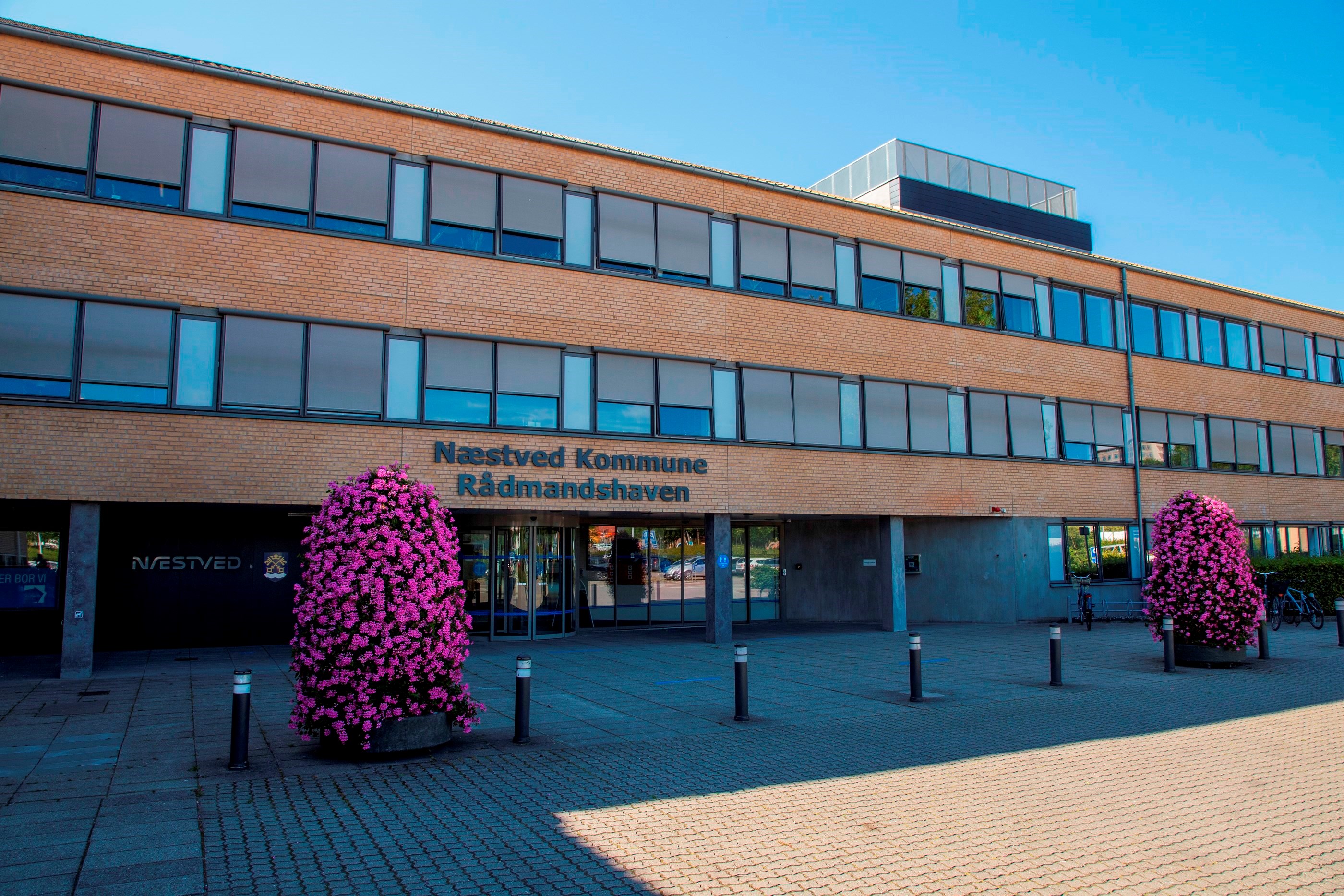 På billedet er der en lang 3 etagers gul murstensbygning, med lange vandrette glaspartier. Foran er der flisebelagt og der er placeret to lilla buske i krukker. På bygningen står der Næstved Kommune Rådmandshaven.