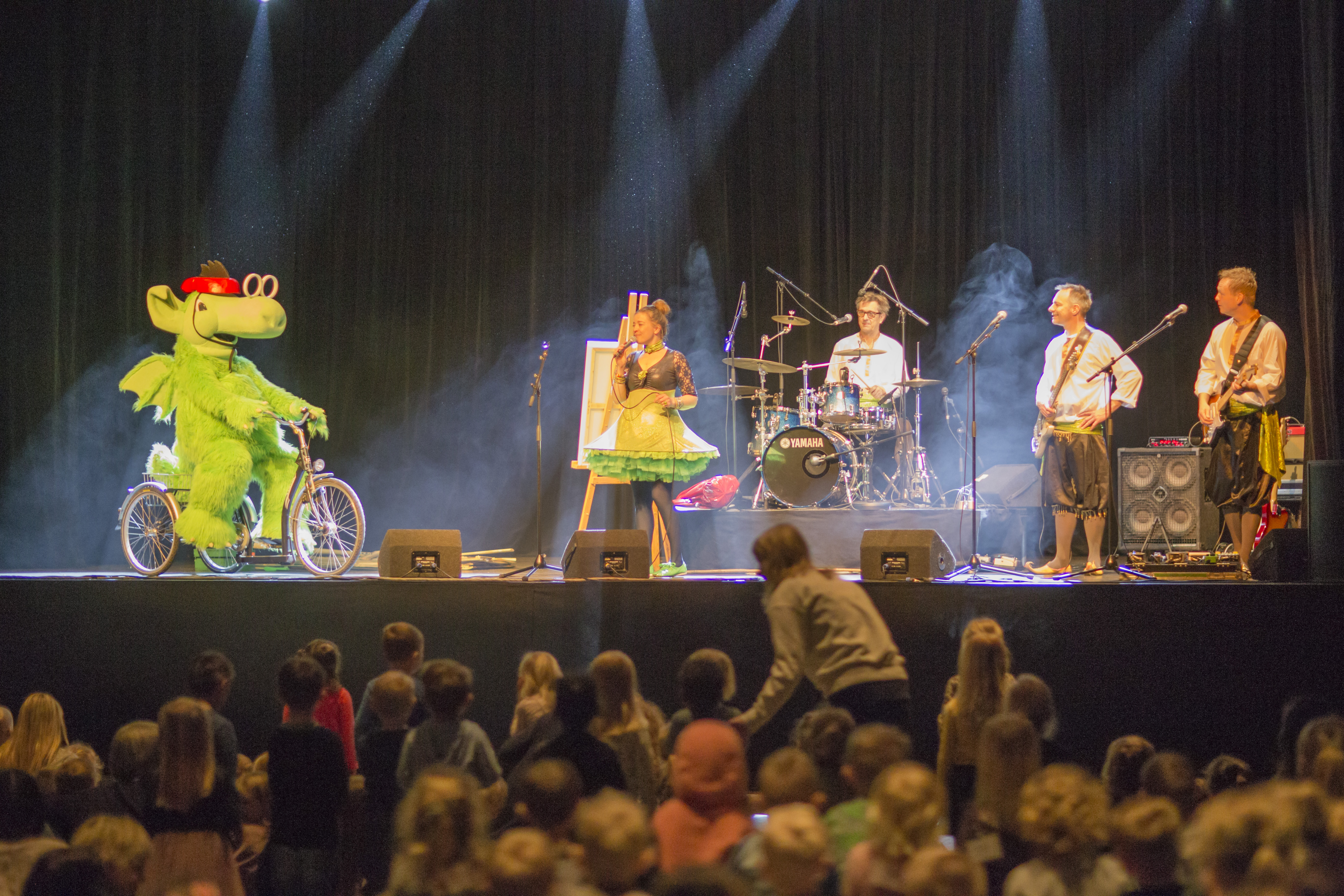 Grøn plysdrage, Næssi, på cykel med rød cykelhjelm og band på scene med børnepublikum i forgrunden. 