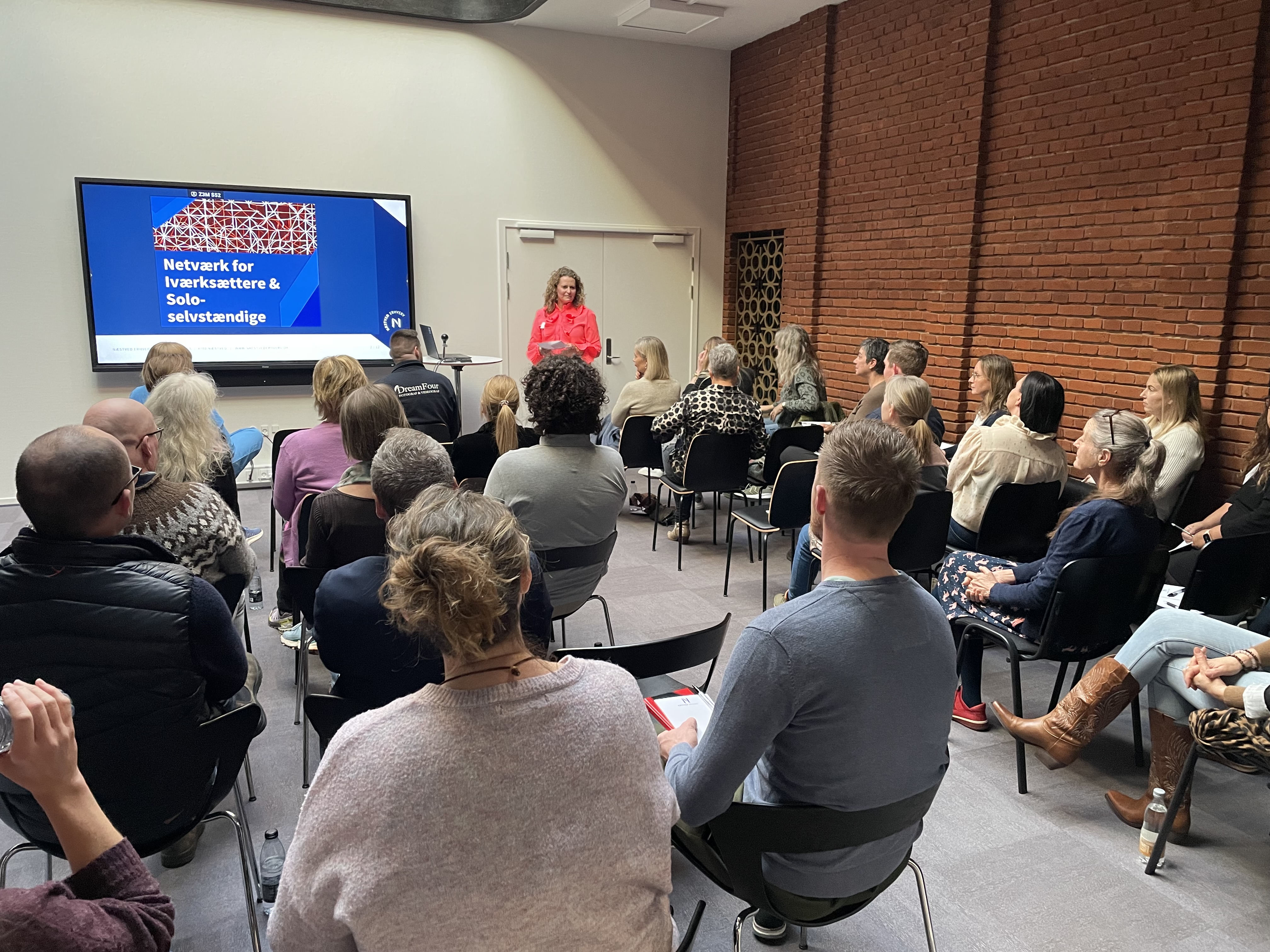 Foredragssituation hvor en kvinde står ved et projektorbillede, mens tilhørerne sidder på stole. 